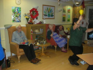 Residents using their musical instruments, whilst staff member dances waving a maraca & tinsel pompom